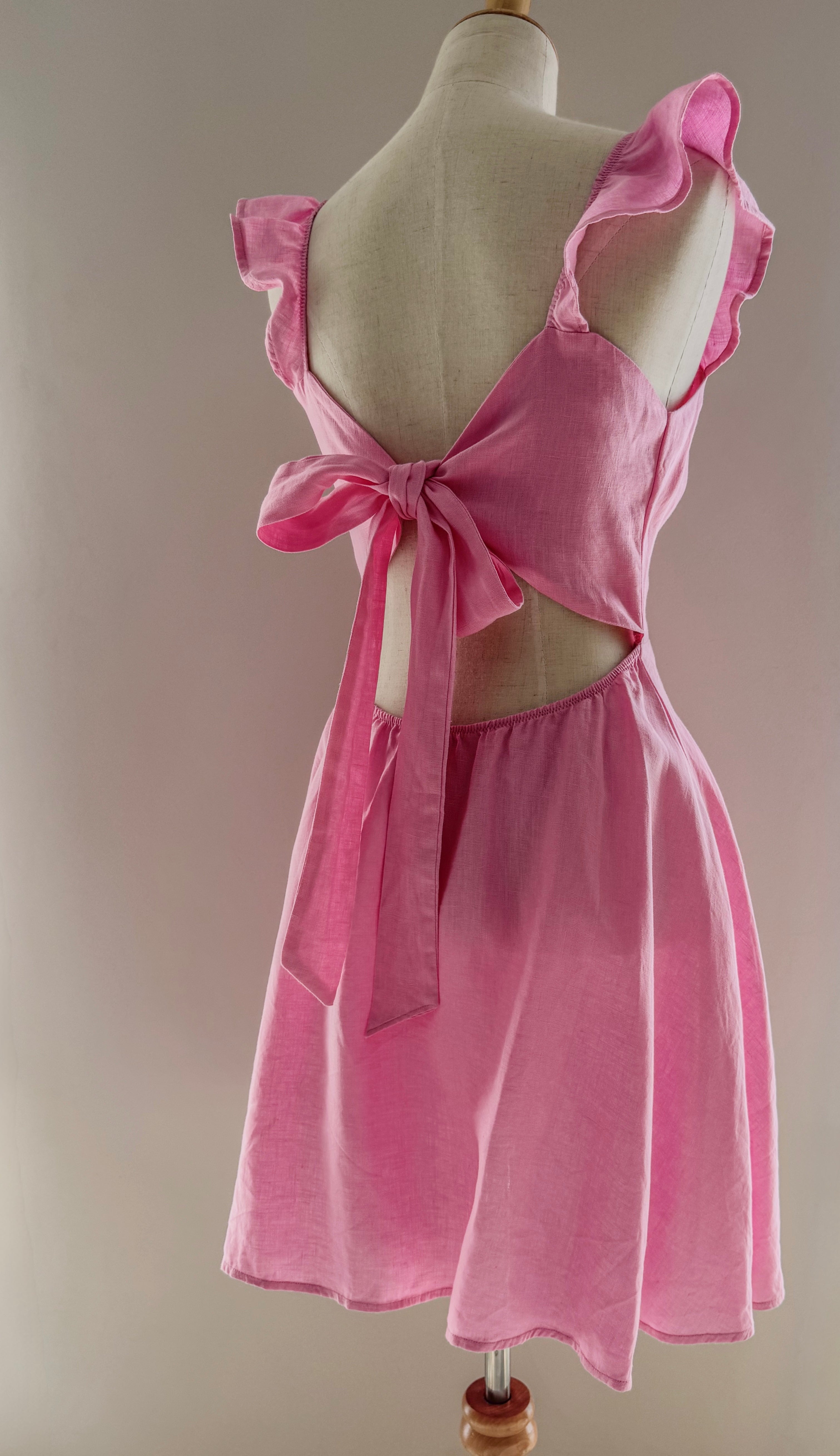 Back view of pink linen dress on a mannequin to showcase the open upper back with bow detail, frilly straps and circle skirt section.