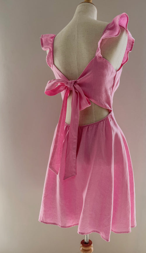 Back view of pink linen dress on a mannequin to showcase the open upper back with bow detail, frilly straps and circle skirt section.