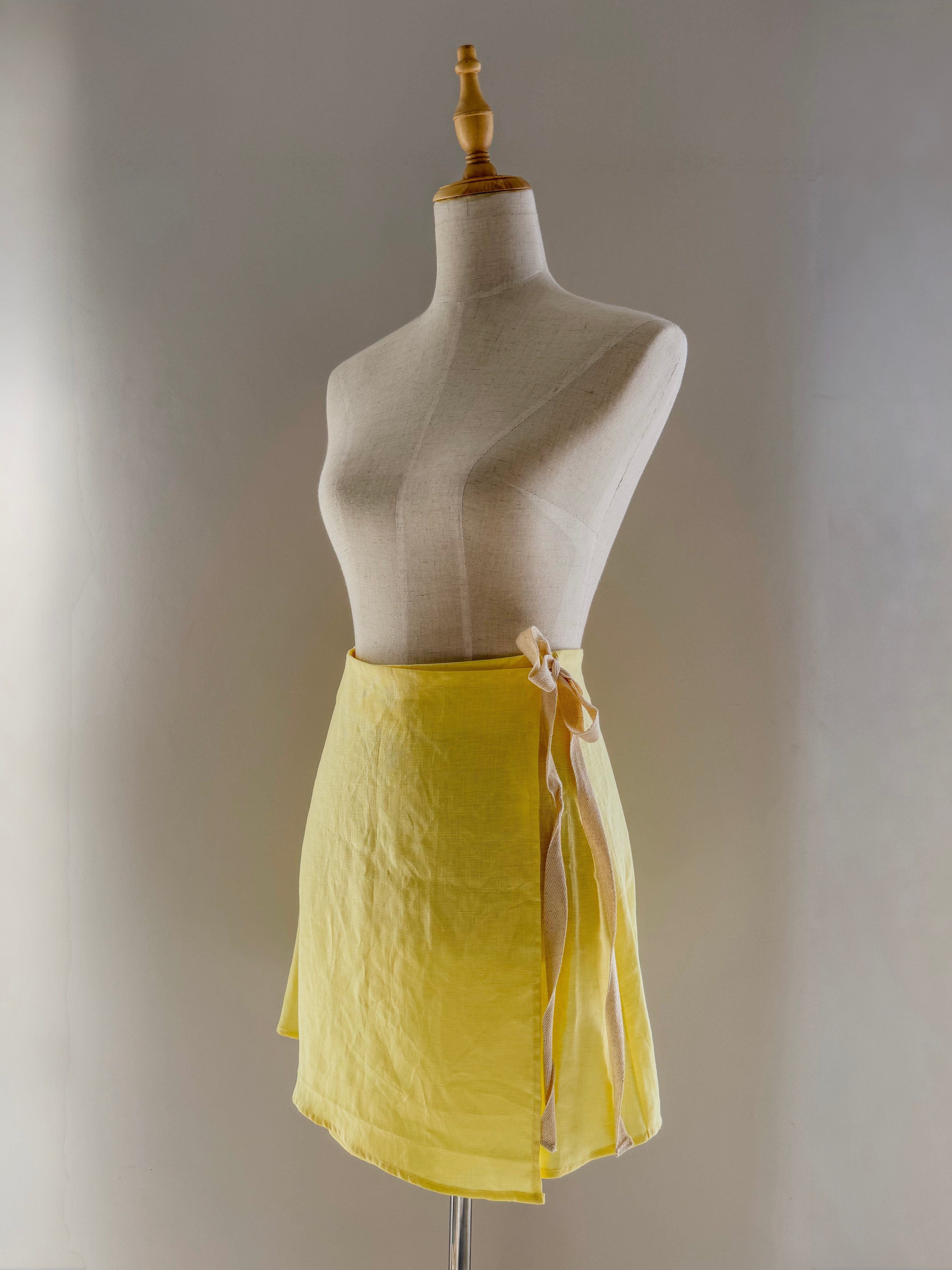 Butter yellow pure linen wrap style high-waisted mini skirt in a more modest length on a mannequin with cotton twill side ties.