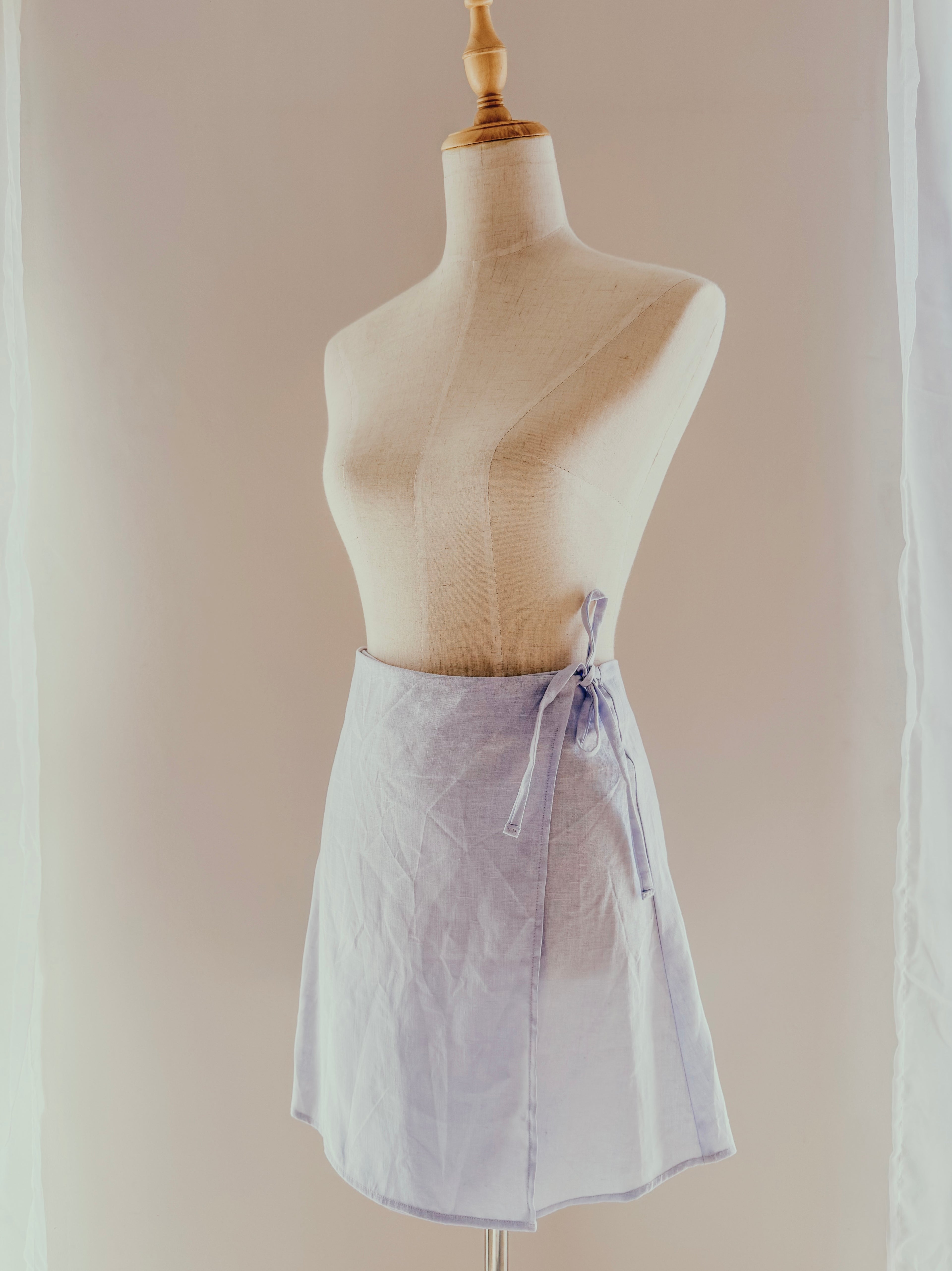 Light blue pure linen wrap style high-waisted mini skirt in a more modest length on a mannequin with side ties.