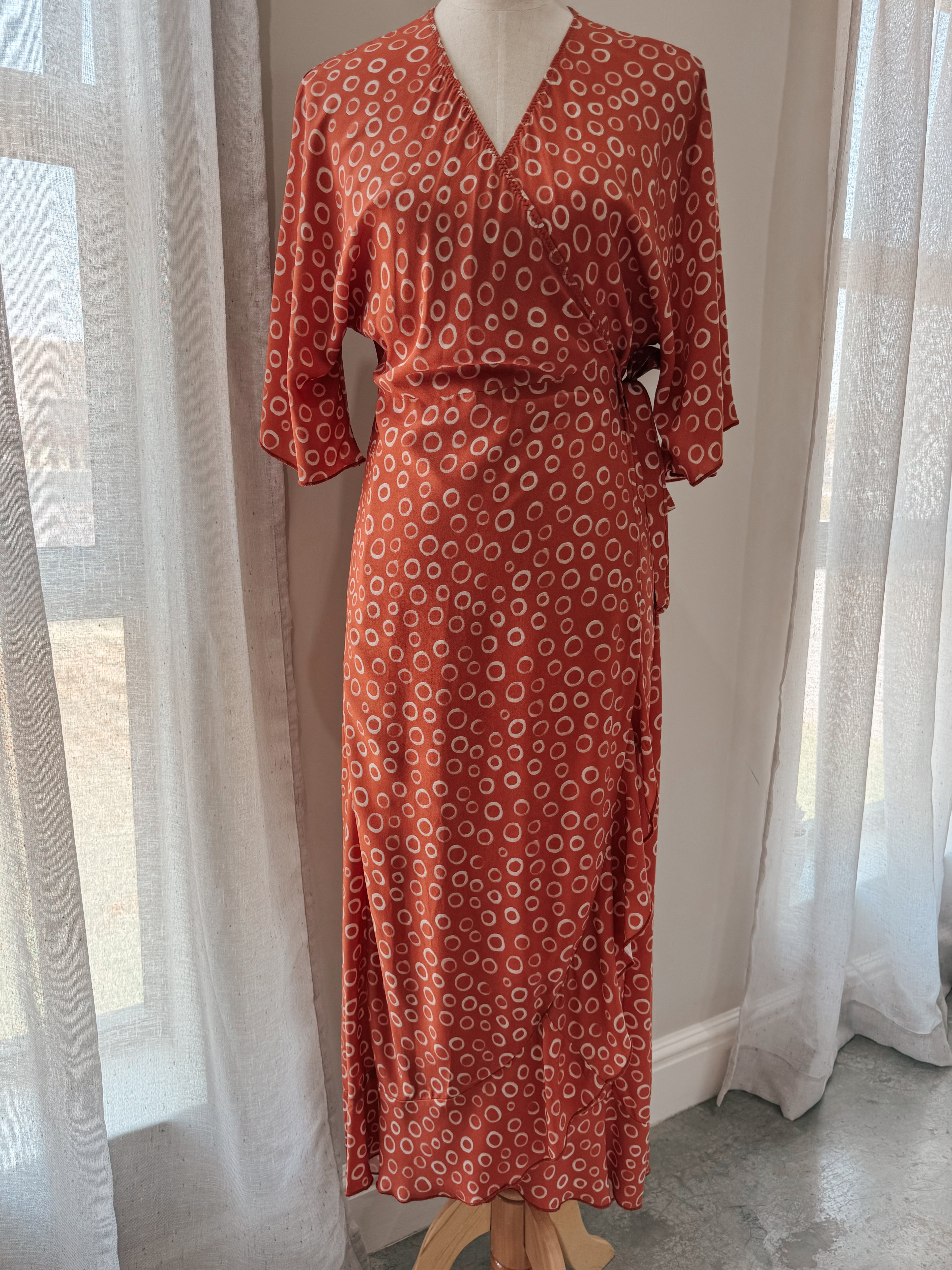 Wrap dress on mannequin made from rusty brown Ecovero Viscose fabric with circular print featuring kimono sleeves and a frilled hem.
