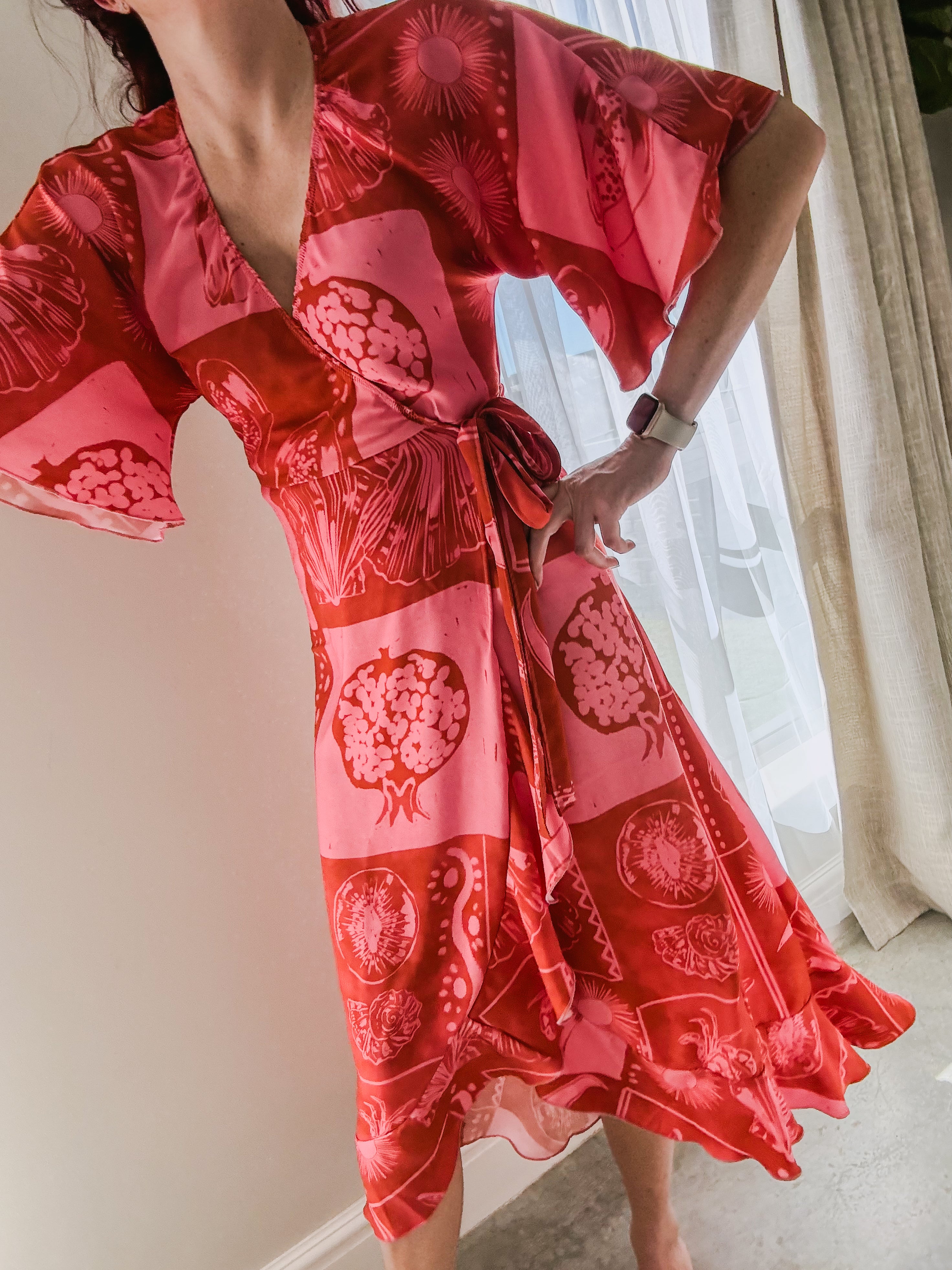Person wearing a red & pink fruit and shells patterned wrap dress made from ecovero viscose, featuring movement in kimono sleeves and frilled hem of bottom.