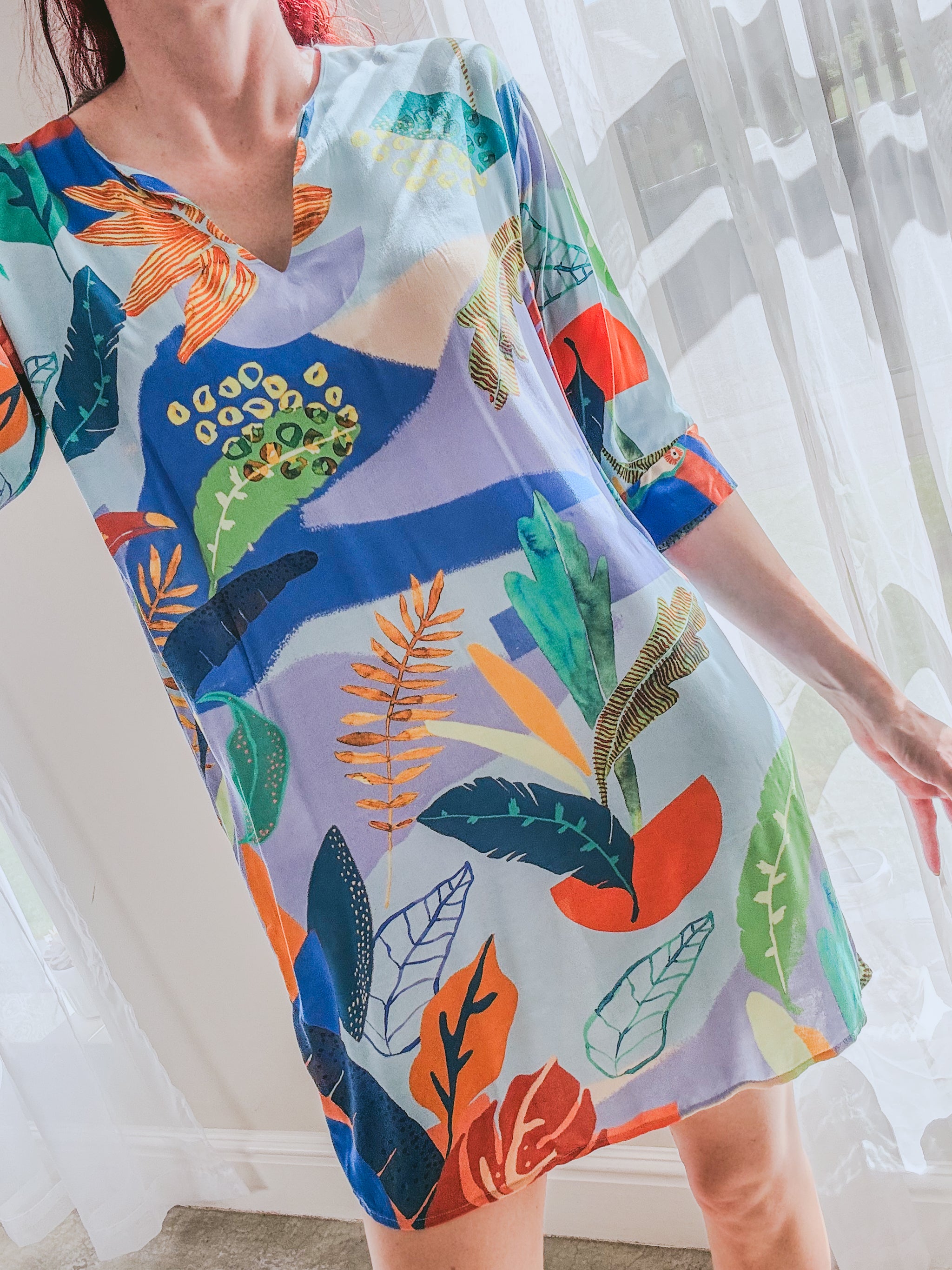 Person wearing a blue above the knee lenght kaftan dress with colourful abstract tropical patterns, featuring a V-slit neckline and 3/4 sleeves with cuff.