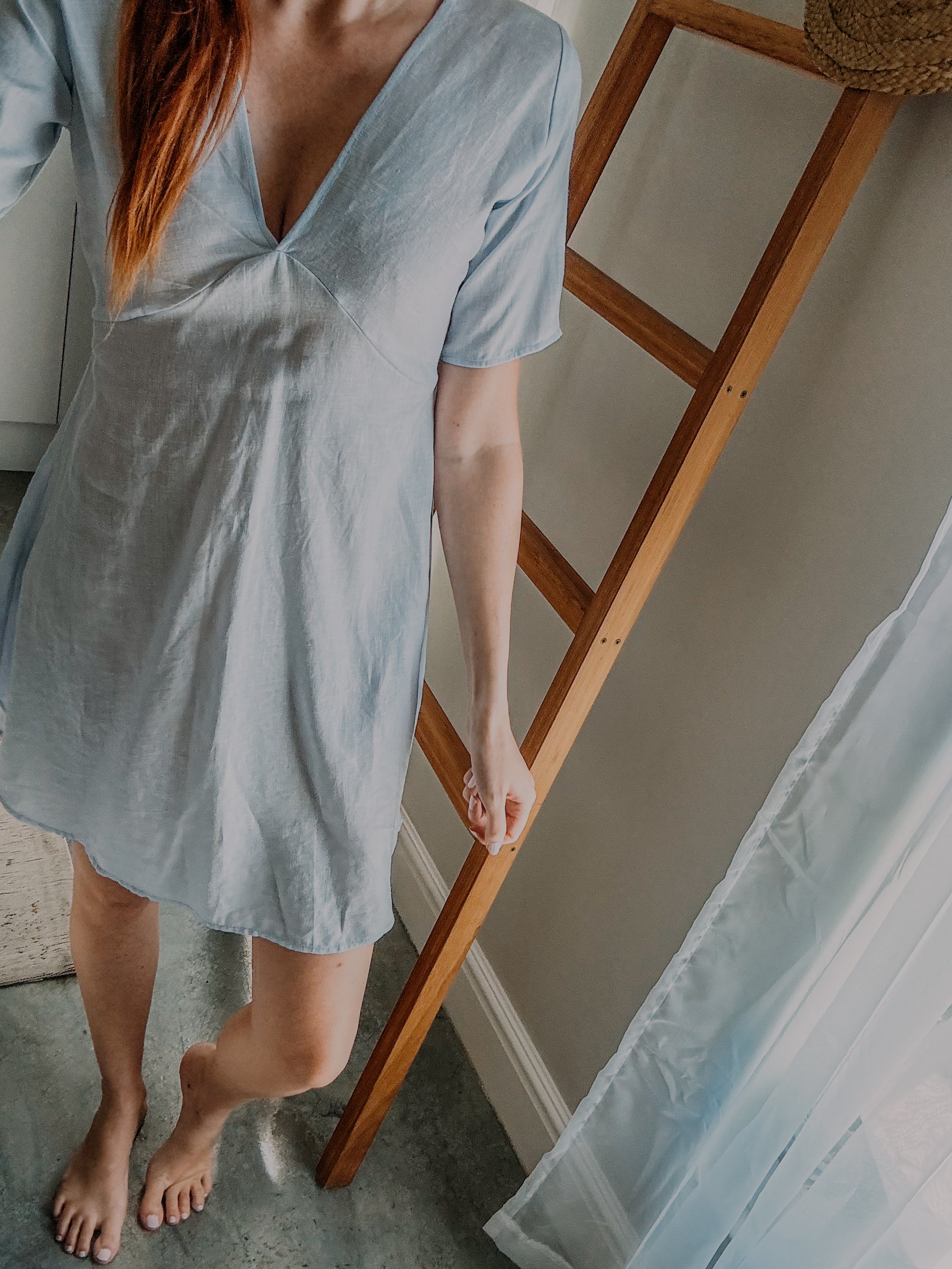 Person wearing a sky blue relaxed pure linen dress with v-neckline and above knee length and above elbow sleeve length.