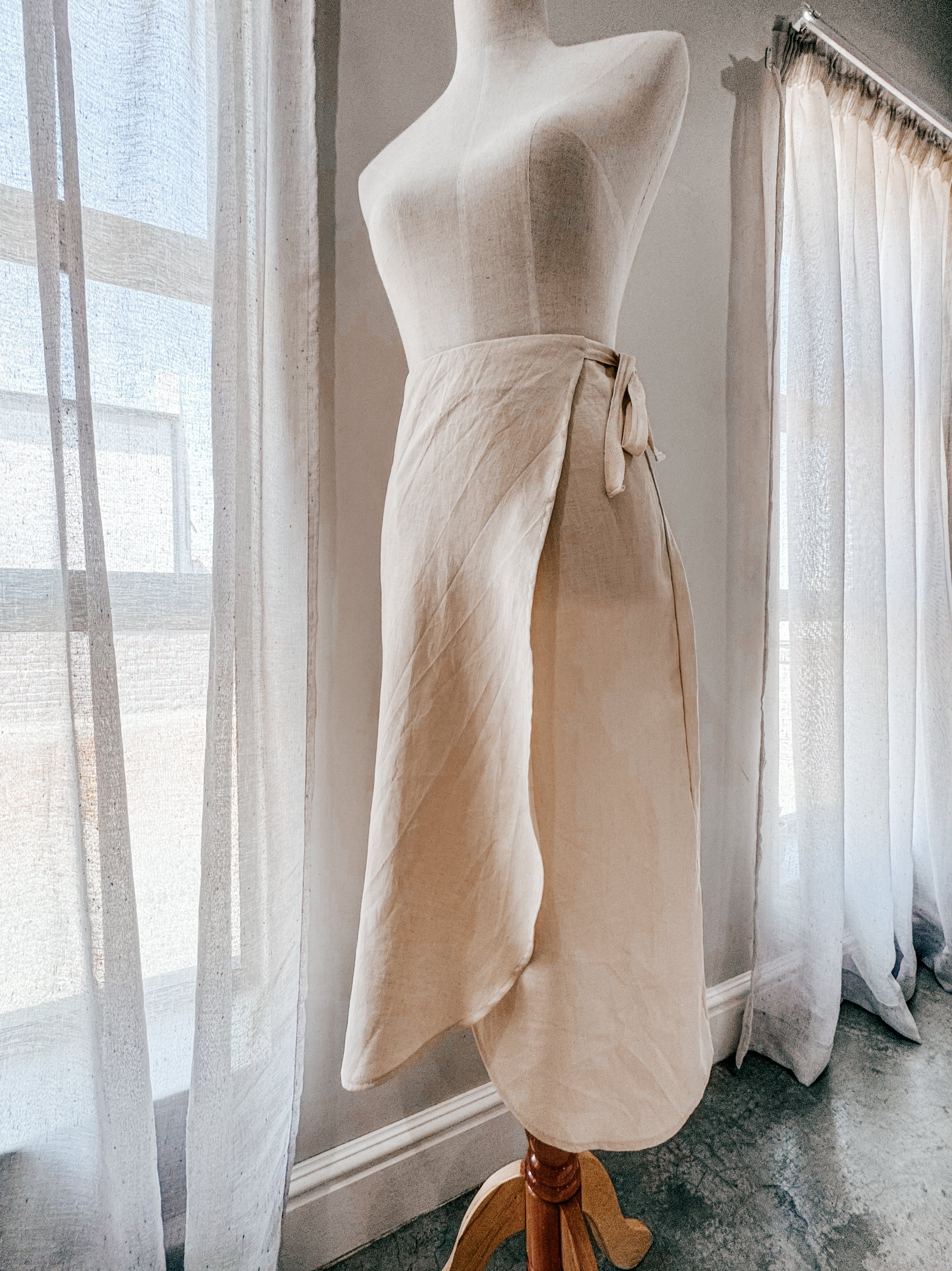 Mannequin featuring a beige high-waisted wrap skirt with curved front hem, made from pure linen with natural twill tape for the side ties.
