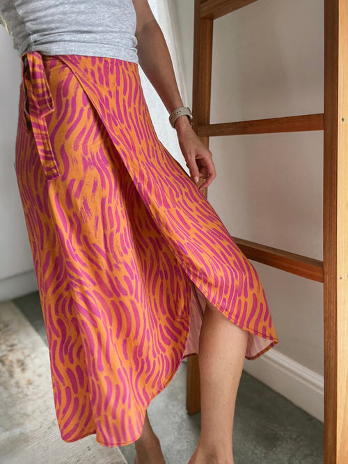 Person wearing a vibrant orange and pink brush stroke patterned midi wrap skirt made from Ecovero viscose.