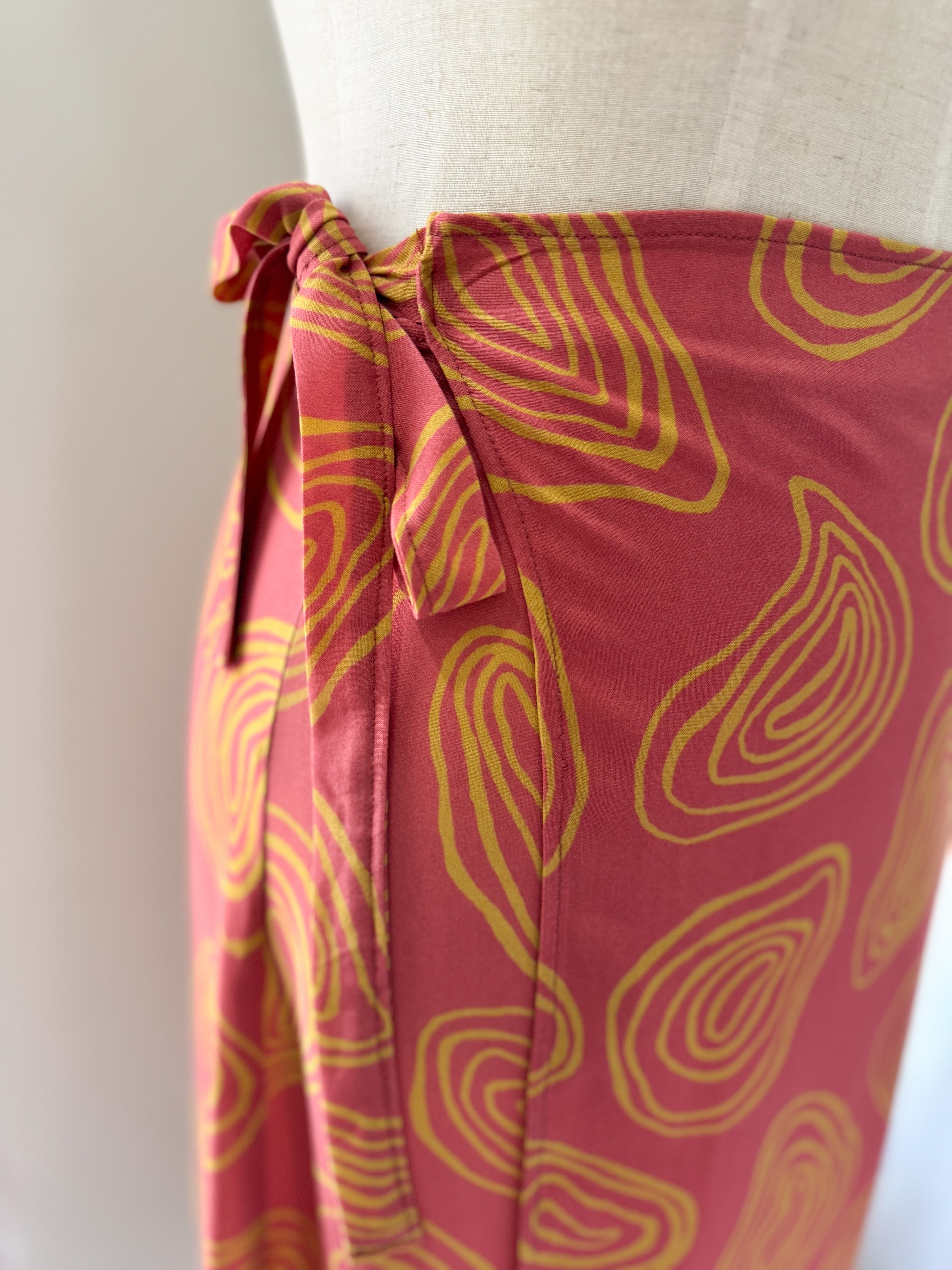 Side view of ties at waist of coral colour high-waisted wrap skirt with yellow squiggle patterns made from Ecovero viscose and displayed on a mannequin.