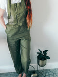 Person wearing olive green dungaree overalls with hand in side pocket.