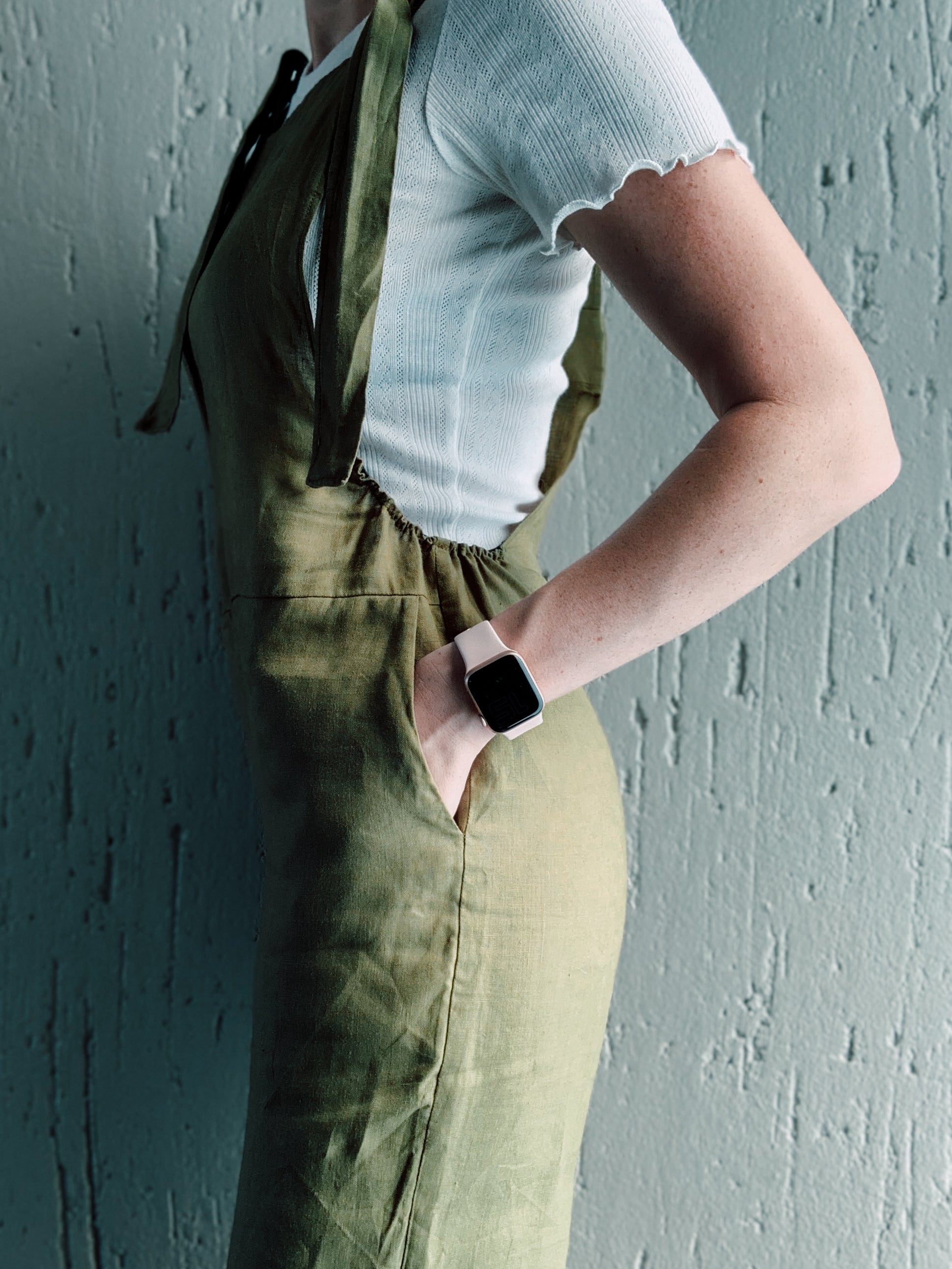 Side view of person wearing olive green dungaree overalls showcasing its practical side pockets and gathered side waist to avoid gaping.