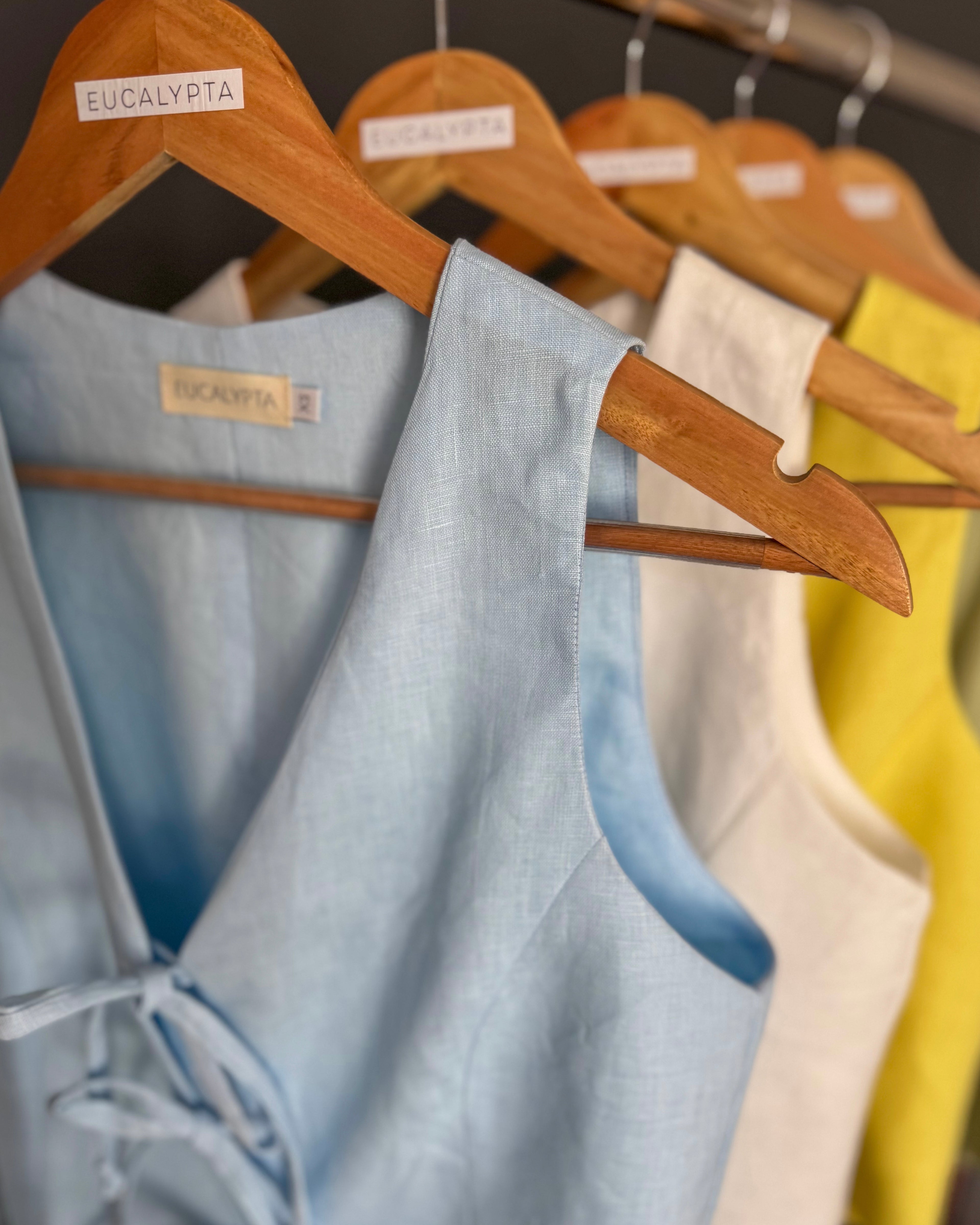 Light blue, white and butter yellow linen waistcoats on a wooden hangers with visible brand label.