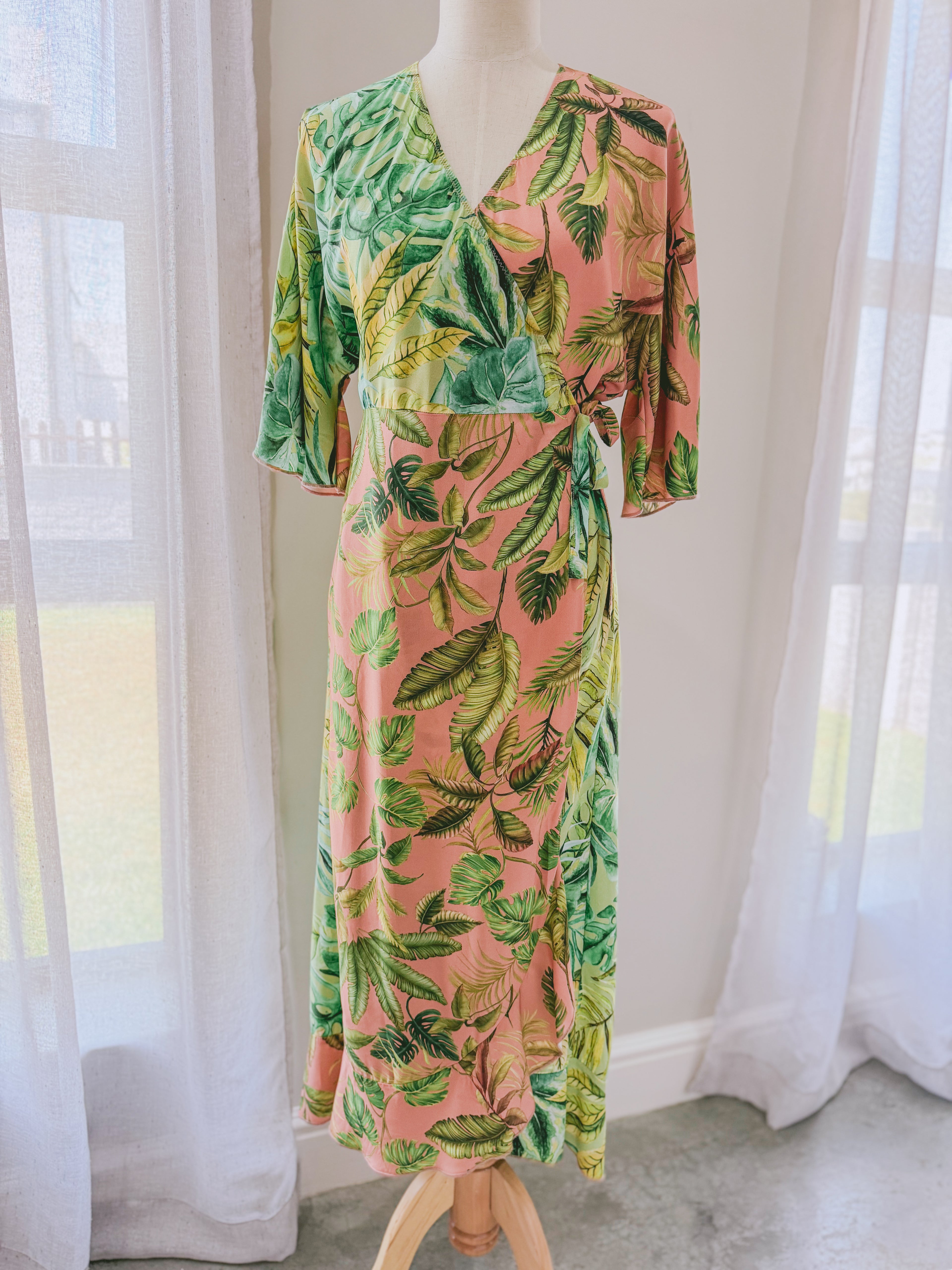 Tropical leaf patterned pink and green Ecovero viscose wrap dress with frilled hem and relaxed sleeves on a mannequin.