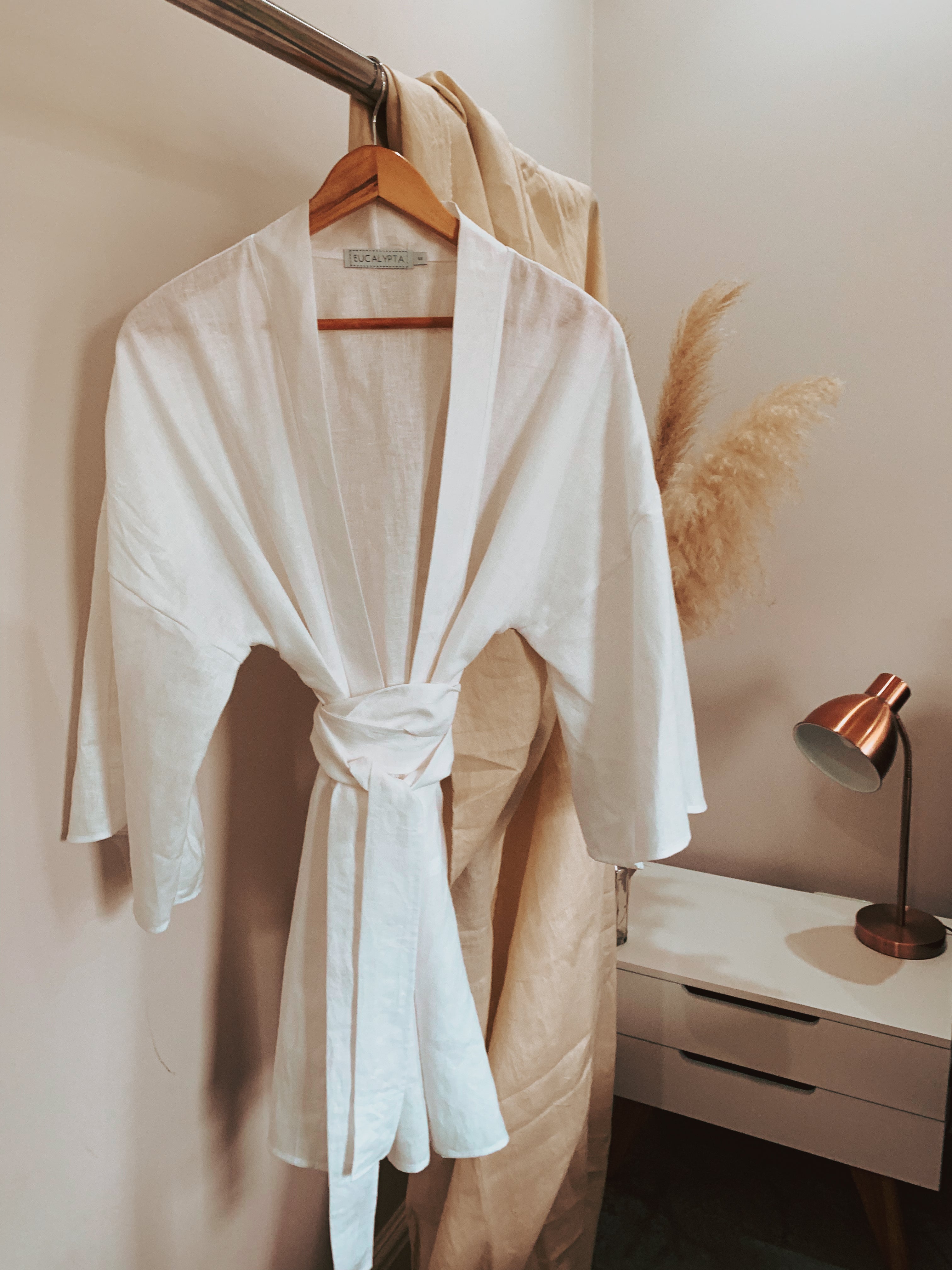 White linen robe hanging on a wooden hanger against a neutral wall with a lamp and decor in the background.