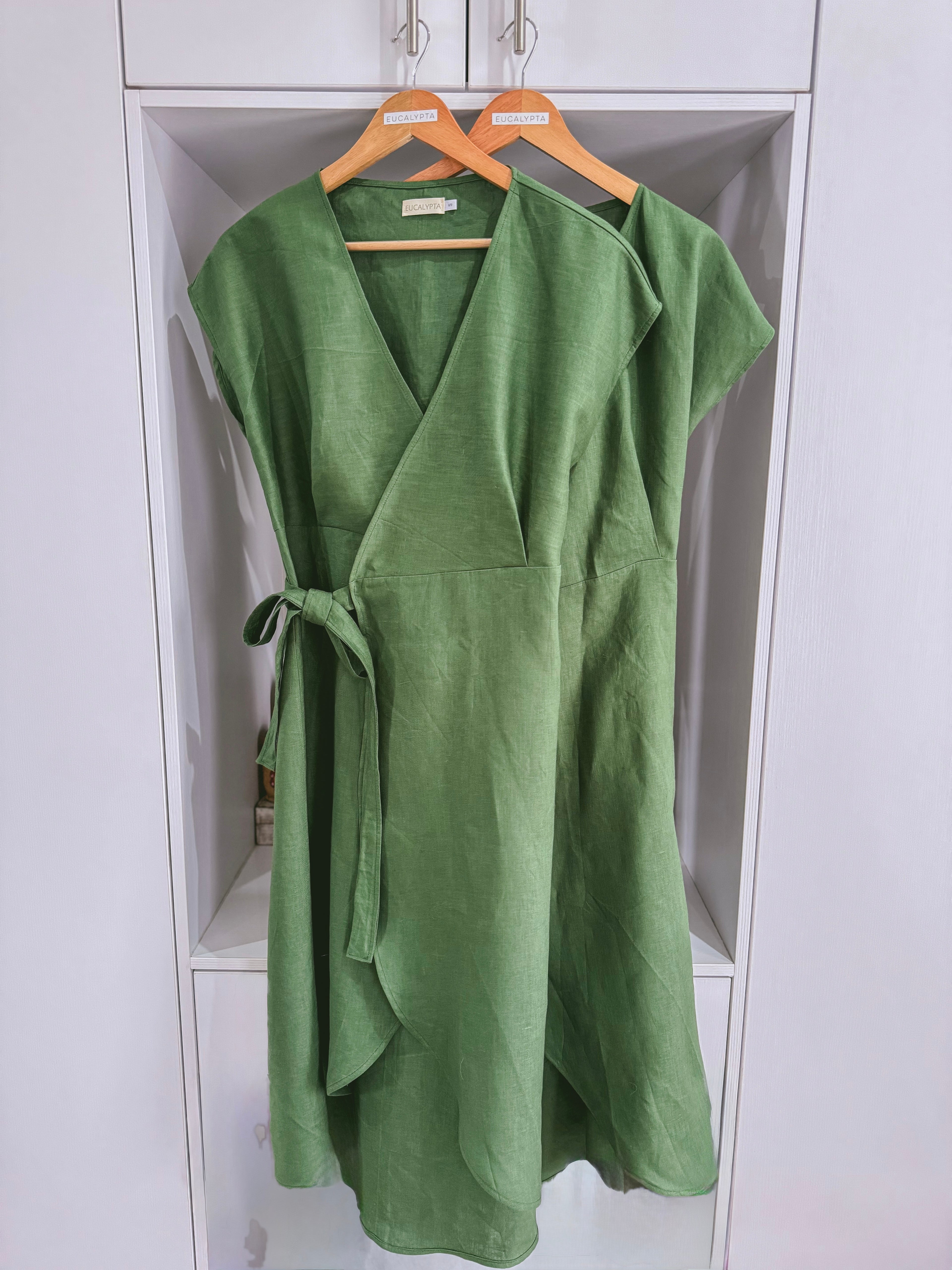 Two green linen wrap dresses hanging on wooden hangers against a white background