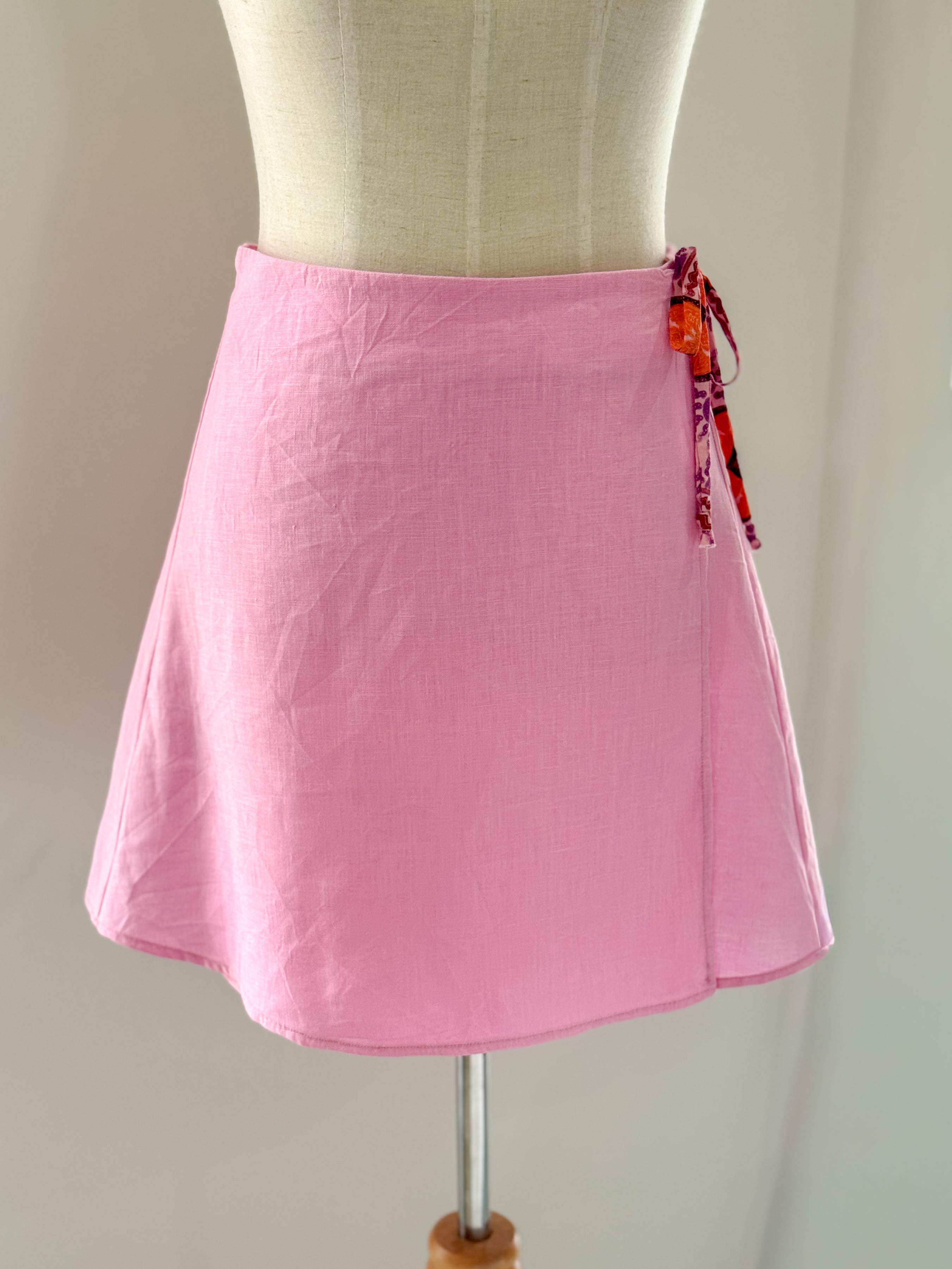 Candy pink pure linen wrap style high-waisted mini skirt in a more modest length on a mannequin with accent ecovero viscose side ties.