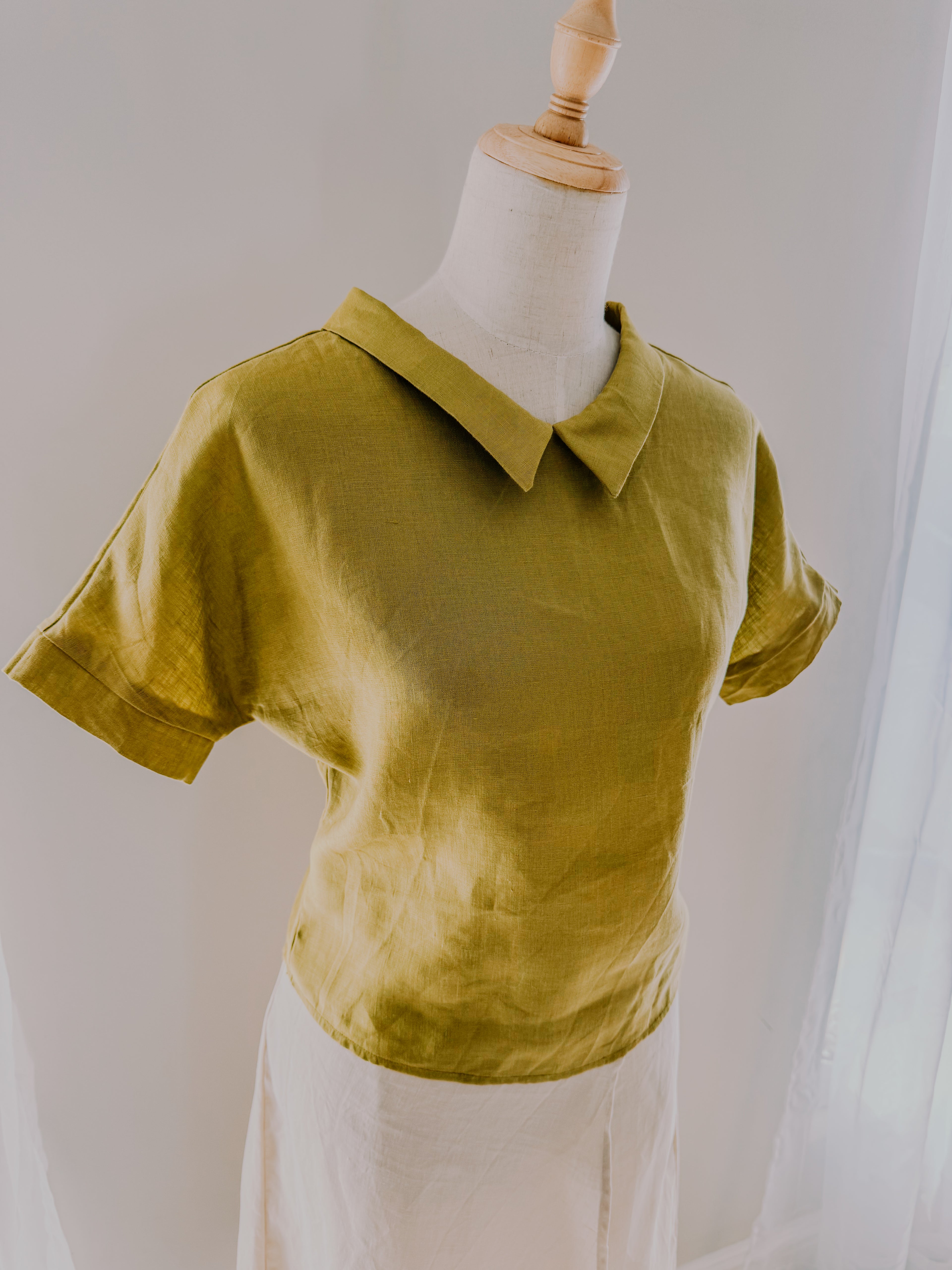 Avocado green short sleeved shirt in relaxed fit on a mannequin with pointed roll collar, grown on sleeve with separate cuff and curved body hem.